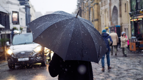 A man walks down a road in Brighton city centre, his back to the camera, carrying an umbrella in heavy rain. Other passers by and a car can be seen outside the entrance to the Brighton Pavilion.