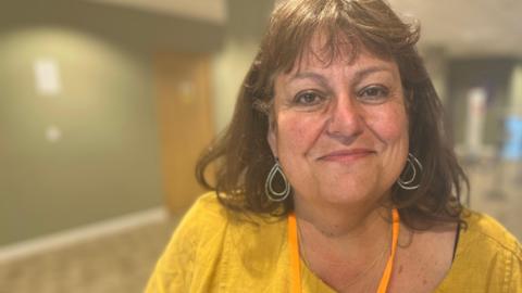 Head and shoulders image of Sarah Conboy in a hallway. She has shoulder length brunette hair, is wearing rounded earrings and a yellow top.