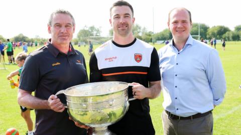 Ulster GAA secretary Ciaran McLaughlin and Armagh football captain Aidan Forker holding the Sam Maguire cup, standing alongside Gordon Lyons at Creggan Gaelic Athletic Club