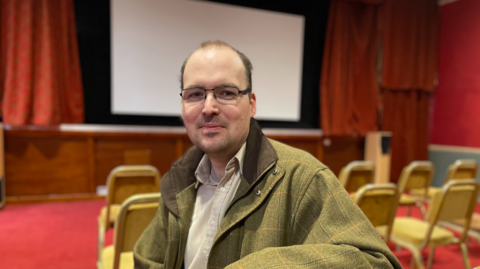 A man with short, balding hair and glasses wearing a green tweed jacket and a neutral shirt. He is sitting on a chair, but has turned to face the camera, which is behind him. There is a large screen and red cinema curtains in the background.