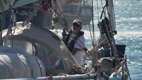 The picture shows a person standing on a sailing boat, wearing a cap and a life jacket. They're positioned near the cockpit area, surrounded by sailing equipment including ropes and sails. In the background, you can see water and a few other boats in the distance.