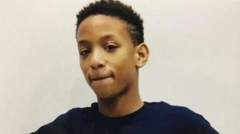 A young teenage boy with brown hair and eyes with an ear piercing looks into the camera against a neutral background.