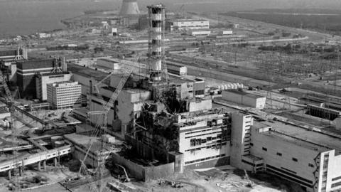 Aerial view of the Chernobyl nuclear power plant's fourth reactor. The center of the image shows a large rectangular concrete structure with extensive damage. Large sections of the roof and upper walls are missing or torn open, exposing internal structural elements. Debris is visible on and around the building.