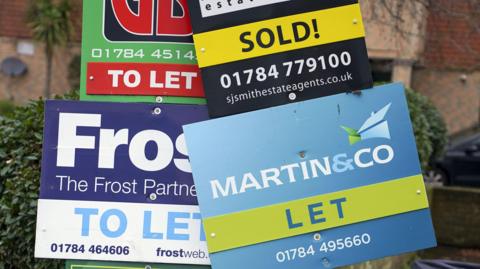 Four 'to let' boards from various different estate agents in close up in front of a blurred hedge and house in the background.