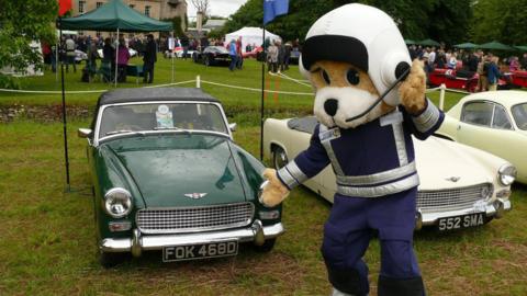 A person wearing a mascot costume of an animal wearing overalls and a white helmet. They are pointing at a classic old car that is parked on the lawn of a large country house.