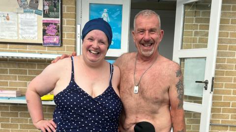 A woman in a polka-dot swimsuit and swimming cap stands next to a man in swimming shorts. He has a stoma bag attached to his stomach and she has one under her costume.