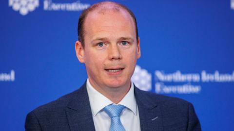 Gordon Lyons - a man with short, dark hair and blue eyes - looks towards the camera during a previous press conference. He is wearing a navy suit, a white shirt and light blue tie. He is standing in front of a blue background with a repeated pattern showing the white logo of the Northern Ireland Executive.