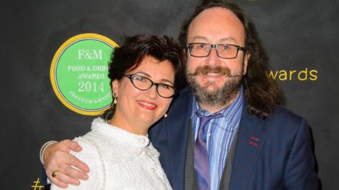 Dave Myers and his wife Lili on the red carpet at an award show. Dave, in a blue suit, has his arm around Lili who is in a white dress. Both are smiling at the camera.