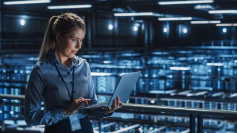 A woman holding a laptop inside a data centre, which has lots of computer systems glowing blue in the background.