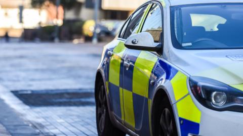 A police car is parked on  a street, with the driver's side in view to the right, and flats behind the car that are blurred out. 