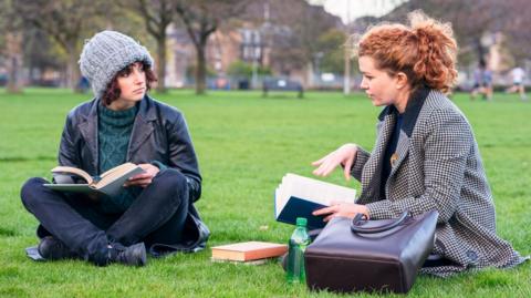 Two female students sitting on grass in conversation. They both have books and are wearing dark clothing