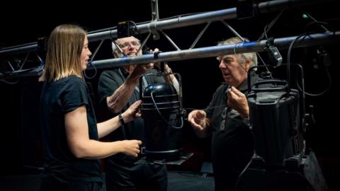 Three people are stood around theatre stage lighting hanging from a rail.