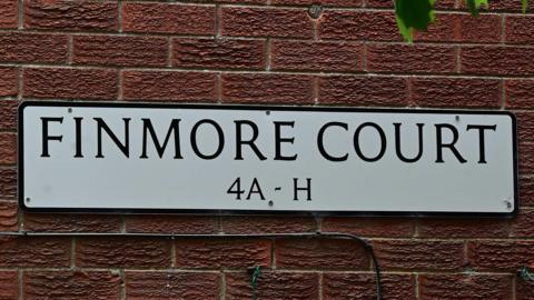 A red brick wall with a white street sign. On the sign, in black letters it says Finmore Court 4A - H 