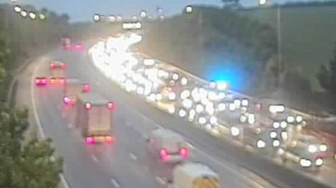 A still taken from a traffic camera looking at the M25. Vehicles are queuing across four lanes on the anti-clockwise carriageway. The blue lights of an emergency vehicle can be seen among the congestion.