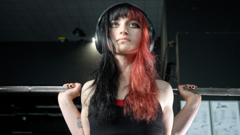 A picture of Lily Churchyard while in the gym. She is pictured from an angle above the camera and looking straight ahead of her. She has a barbell resting over her shoulders ready for her to perform a squat. She is wearing black headphones over her ears. She has long hair, with one half which is black and the other half red. She is wearing a black top and she has dark eye shadow on with a nose ring.