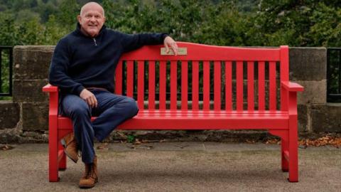 Dean Dyble sits on a bright red wooden red bench. He is an older man, with a bald head and a blue jumper, blue jeans and brown boots. In the background, trees and a stone wall can be seen.