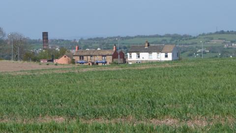 Rural landscape with open fields leading towards a group of houses in the distance with hills in the far distance