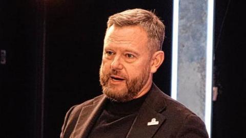 Mark Ashton has short brown hair and is wearing a brown suit jacket over a black rollneck top. He is speaking on stage.