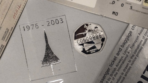 A shiny 50p piece which says "CONCORDE" on with a design of the aircraft underneath is on a table surrounded by Concorde-related documents, including one in silver which says "1976-2003"
