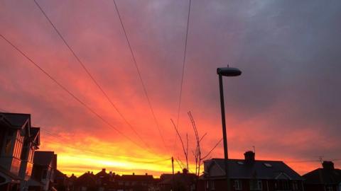 A street lamp is in front of a red sky. The roofs of houses and phone lines can also be seen against the sunset.
