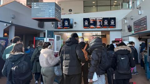 Dozens of pasengers wait in the station lobby with screens above on a wall showing train information.