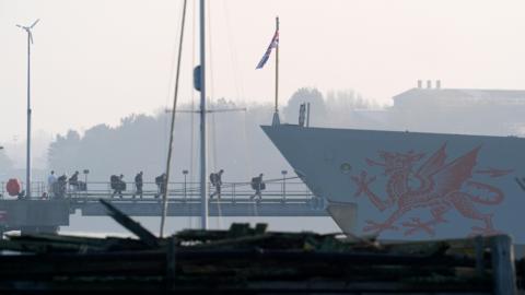 Crew members board the HMS Dragon during ammunitioning operations. A red dragon is painted on the side of the grey ship.