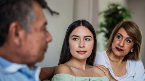 A father, daughter in her 20s and mother sitting on a sofa together. They have a serious looking expression on their faces