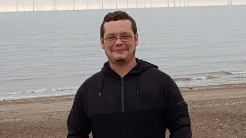 A photo of Aaron Keightley wearing a black hoody. He is standing on a beach with the water behind. 