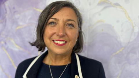 A woman with short dark coloured hair is looking directly at the camera and smiling. She has blue eyes and red lipstick. She is wearing a black blazer with a white trim and silver necklace. She is standing infront of a light purple and white backdrop. 