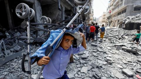 A Palestinian boy carries a stroller with debris surrounding him