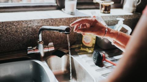 A kitchen tap running with a thin stream of water, in the background there is a silver steel draining board with a washing up brush on it, a bottle of washing up liquid and a granite effect worktop seen in the background.