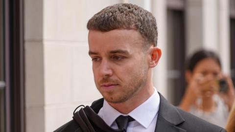 A man wearing a suit and tie walking into court, background blurred but woman can be seen taking a photo.