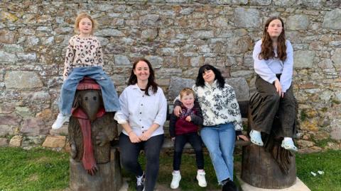 A family photo with two young girls, two women and a young boy sitting on a wooden bench. 