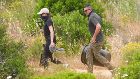 Search officers walk through an area of shrubland holding a spade, a bin and a plastic bag.