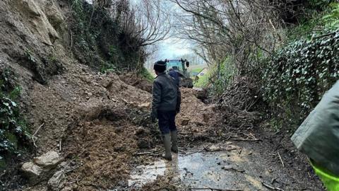 A man surveys a landslip across a road with a tractor in the background.