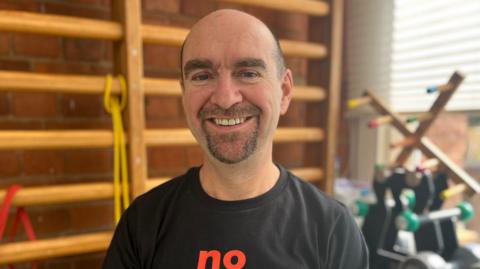 The head and shoulders of a bald man wearing a black t-shirt can be seen in front of a gym setting. He is in his early fifties, has a beard and is smiling.