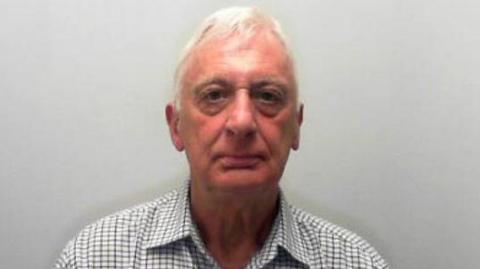 Man with white hair wearing a checked shirt looks into a camera.
