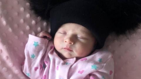 A sleeping baby wearing a pink baby grow with blue stars and pink hearts on it. She is on a pink blanket and wearing a black fluffy hat