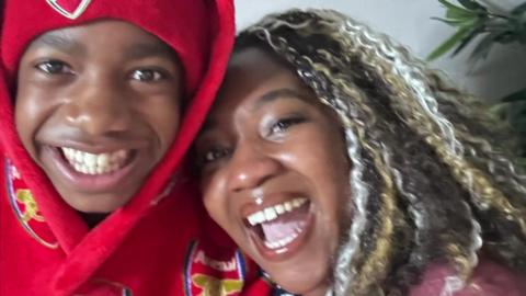 A young boy wears a red Arsenal hoodie and hat, next to a woman with multi-coloured hair. Both are smiling. 
