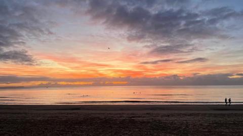 The sun is just rising over the sea creating an orange glow in the sky. The beach itself is still in darkness and two lone joggers are about to run out of the picture. 