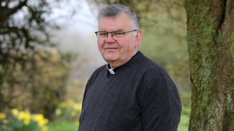 Mark Christian is pictured wearing a dark grey V-neck jumper with a white dog collar. He is standing outside, next to a tree, on a cloudy day. He has short grey hair and is wearing black-rimmed glasses. He is looking at the camera in a slightly pensive way.