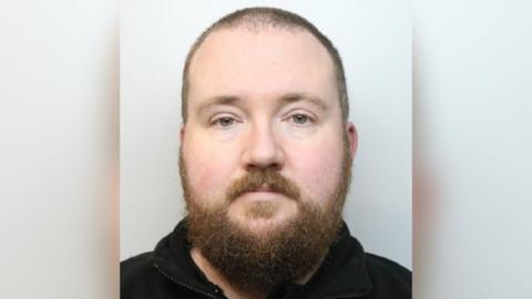 A police photo shows a man with a large beard and wearing a dark fleece