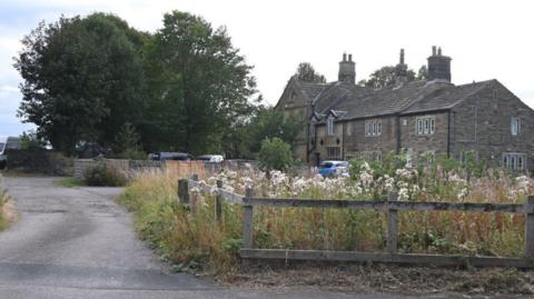A large stone building set back from the roadside, bordered by a wooden fence, greenery and trees.