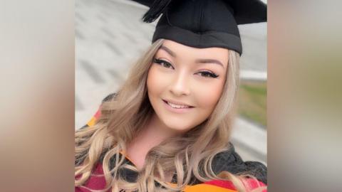 A woman with long blonde hair, a graduation cap and yellow, red and black graduation robe, takes a selfie.