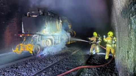 A maintenance vehicle in Standedge Tunnel being hosed down by firefighters wearing protective clothing 