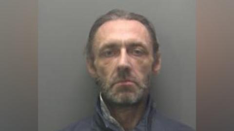 A police mugshot of James Conroy. He has pulled-back brown hair and a greying beard. He is wearing a dark grey anorak done up to his neck. 