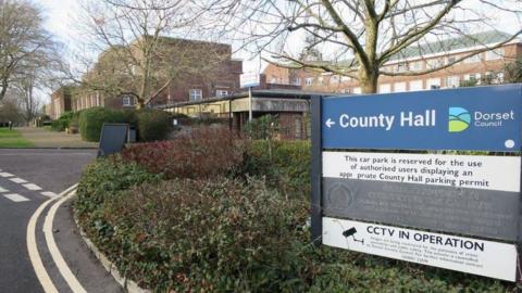 County Hall in Dorchester. In the foreground is a signpost to the large redbrick office building which is surrounded by shrubs.