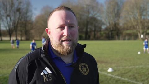 Nick Stephens in a black tracksuit top with club badge, standing in front of training pitch with kids playing in the background
