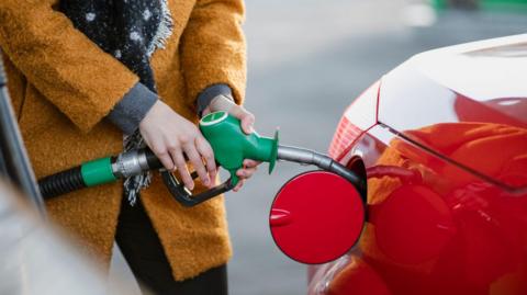 Close up of a woman in an ochre coloured warm coat inserting a petrol nozzle into a bright red car petrol tank.