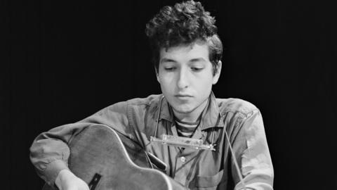 Black and white image of a young Bob Dylan in 1962. He has a dark quiff of hair and wears a buttoned shirt. A harmonica is strapped around his neck and an acoustic guitar in his hands.